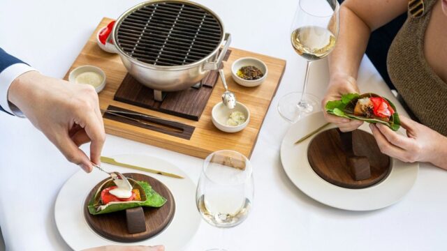 Twopeopleatadiningtablewithglassesofwineservingthemselvesfoodoffofatabletopgrill